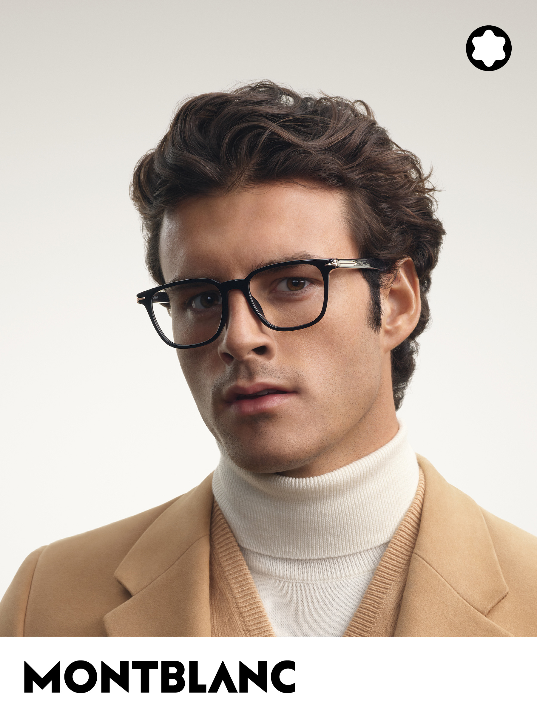 Un homme aux cheveux bouclés et portant des lunettes, vêtu d'un manteau beige et d'un pull à col roulé, devant un fond neutre. On aperçoit le logo Montblanc.
