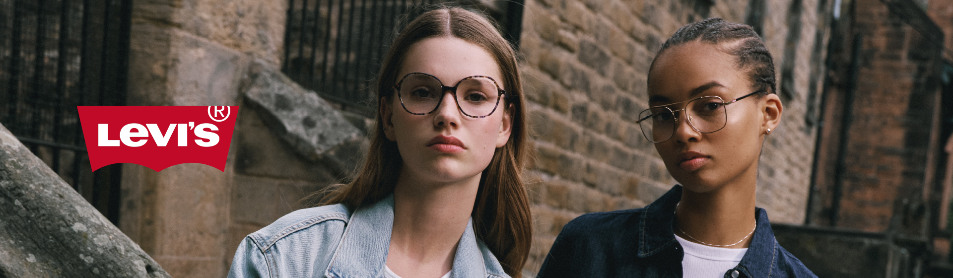 Dwie osoby w okularach i dżinsowych kurtkach stoją przed kamiennym murem. Z boku widoczne jest logo Levi's.