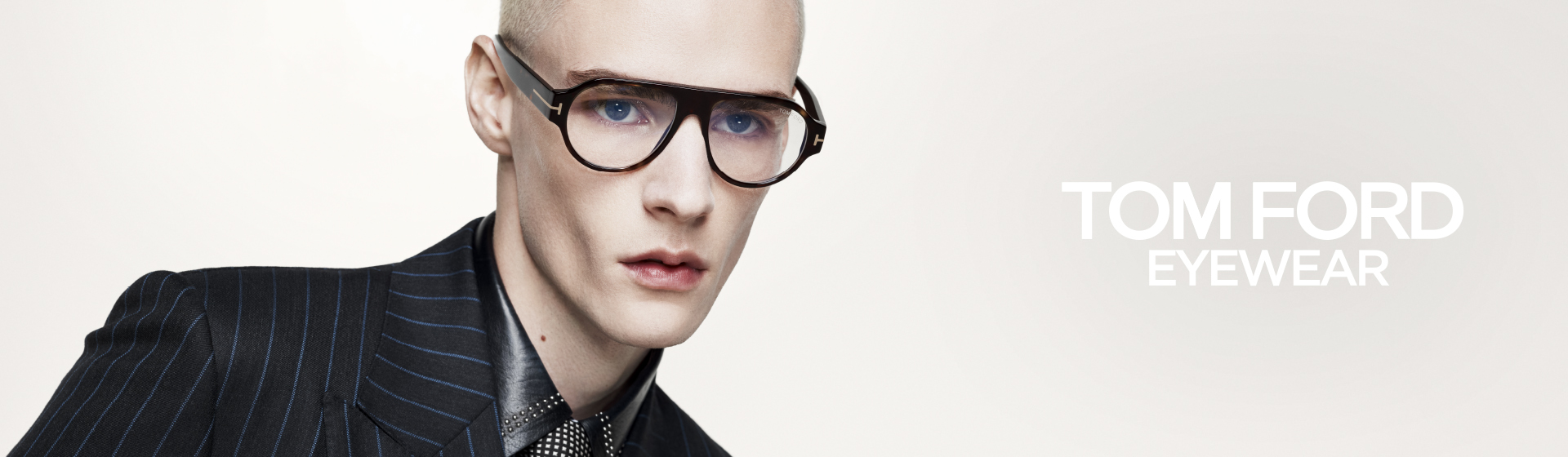 Eine Person mit einer modischen Brille, rasiertem Kopf und einem gestreiften Anzug vor einem hellen Hintergrund mit dem Schriftzug „Tom Ford Eyewear“.