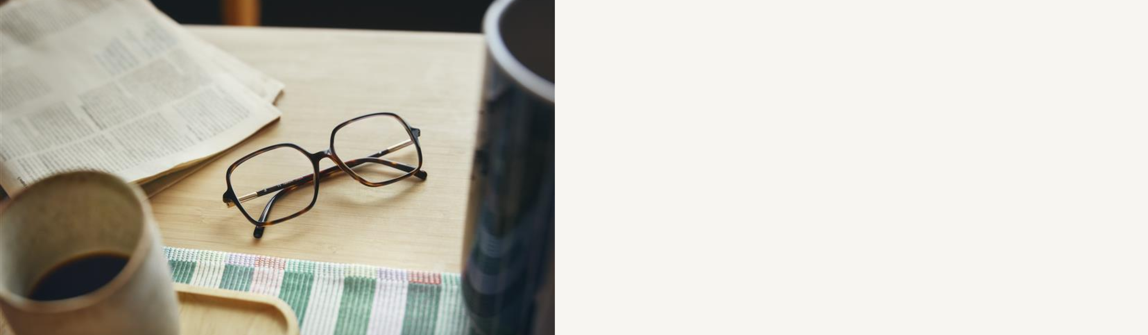 Eine Brille liegt auf einem Holztisch neben einer aufgeschlagenen Zeitung, einer Tasse Kaffee und einem gestreiften Tuch.