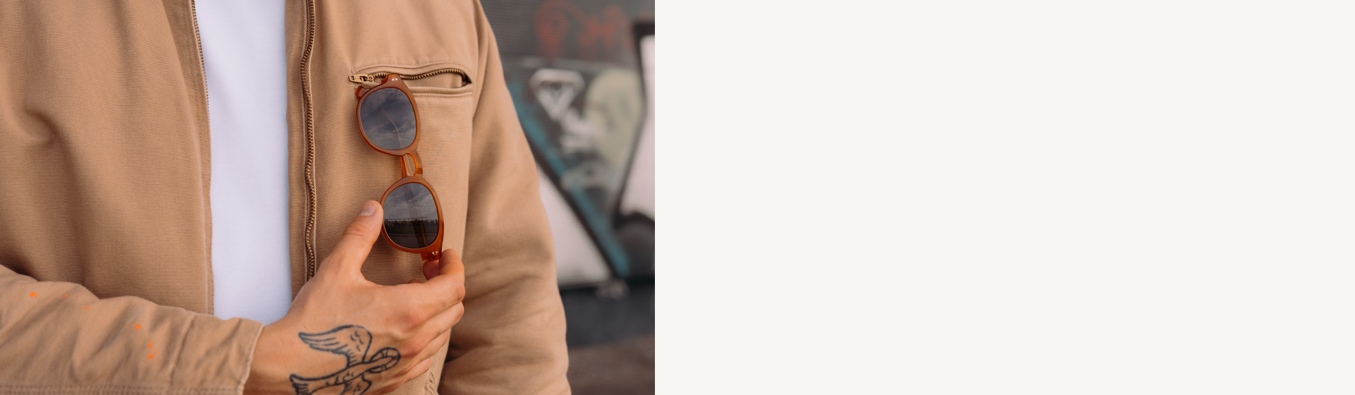 Mano con un tatuaggio di un uccello che tiene degli occhiali da sole rotondi marroni attaccati alla tasca con cerniera di una giacca marrone chiaro.