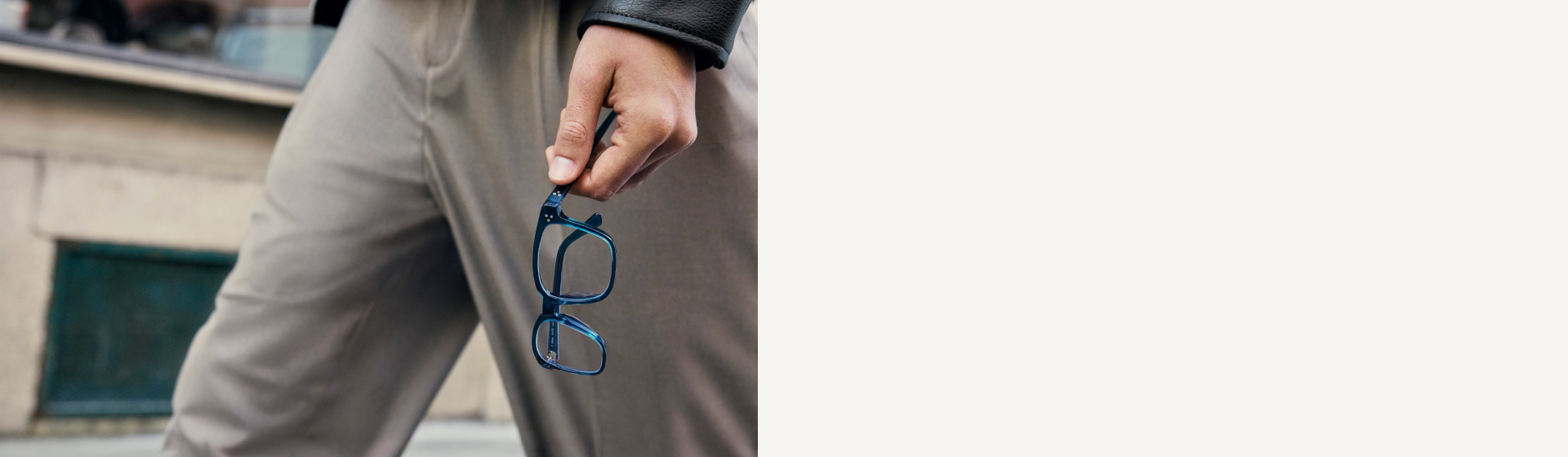 Mano che tiene un paio di occhiali blu sulla stanghetta, giacca di pelle e pantaloni grigi sullo sfondo.