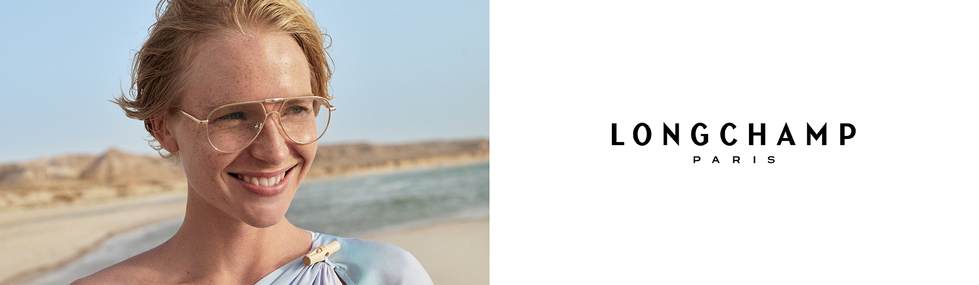 Uśmiechnięta osoba w okularach na plaży, z krótkimi włosami, ubrana w jasnoniebieską bluzkę. Widoczne jest logo Longchamp Paris.
