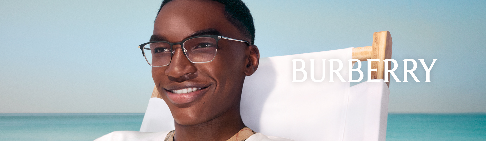 Une personne souriante portant des lunettes et un haut blanc avec le logo Burberry, devant un océan calme.