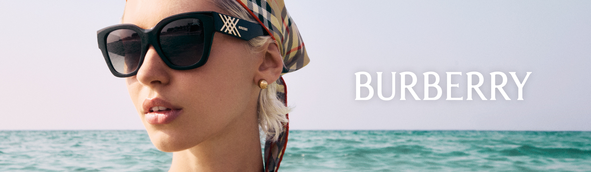 Kobieta w okularach przeciwsłonecznych i chuście w wzór Burberry stoi na plaży, a w tle widać morze.