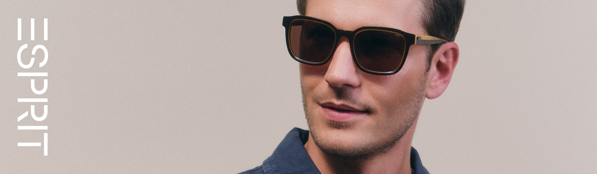 Mężczyzna o brązowych włosach, w ciemnych okularach przeciwsłonecznych i niebieskiej koszuli, stojący na beżowym tle. Napis „Esprit” umieszczony z boku.