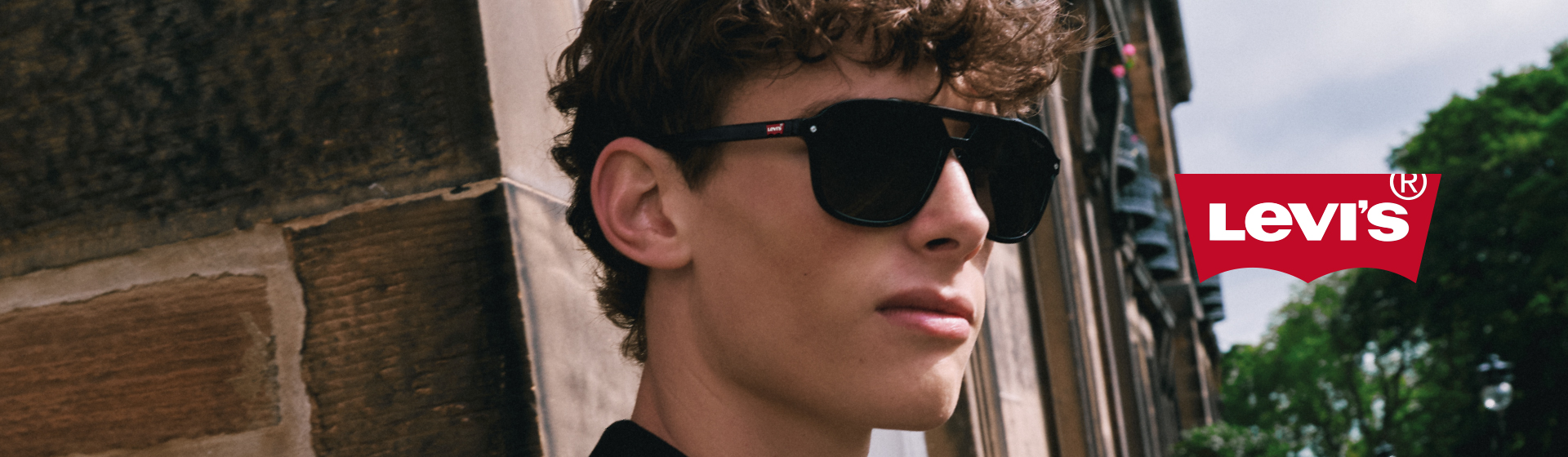 Person mit Sonnenbrille und schwarzem Hemd, die sich neben einem Levi's-Logo an eine Wand lehnt, vor einem urbanen Hintergrund.