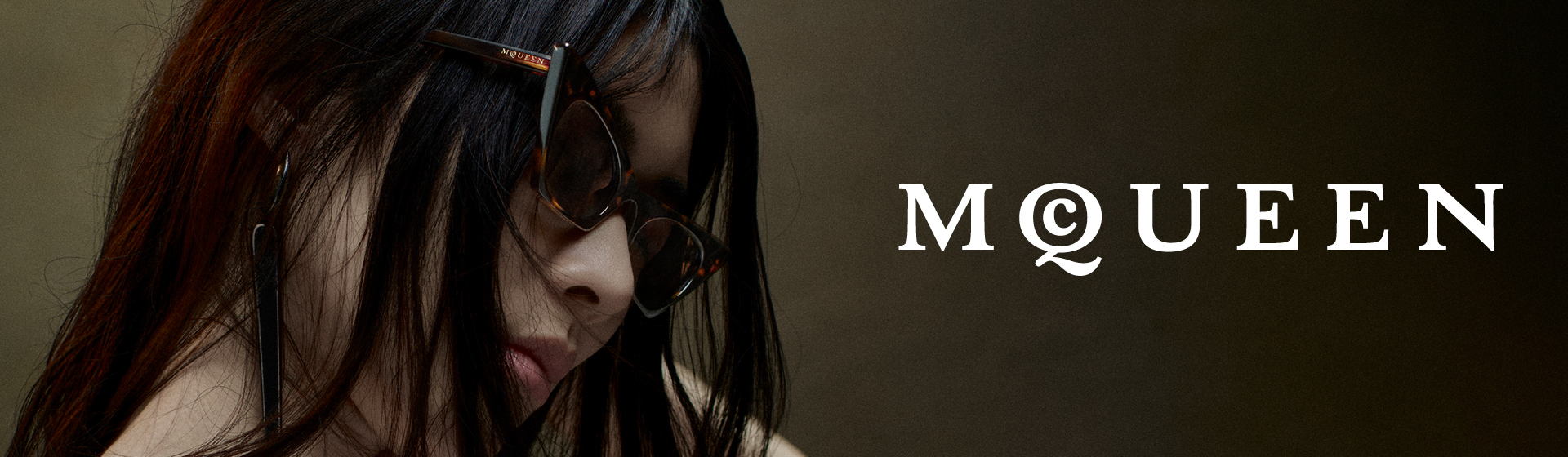 Osoba z długimi włosami i okularami przeciwsłonecznymi na tle brązowego tła. Słowo „McQueen” napisane jest pogrubionymi białymi literami.