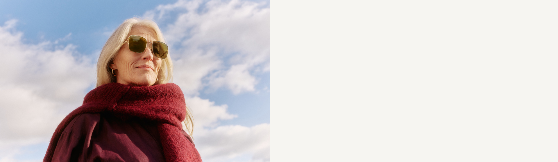 Silberhaarige Frau mit Sonnenbrille und einem kuscheligen burgunderroten Schal, die gegen den blauen Himmel nach oben blickt.