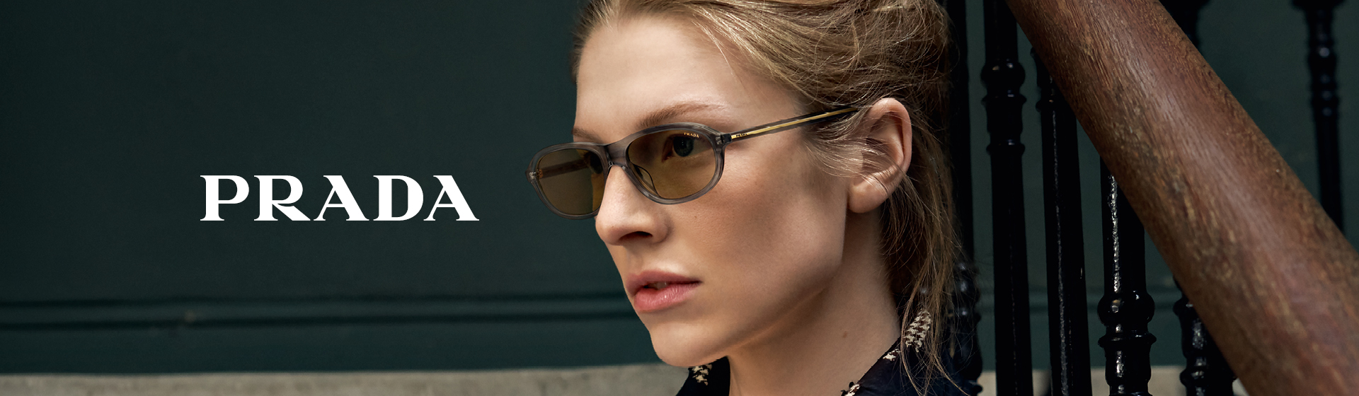 Eine Person mit Sonnenbrille und schwarzer Kleidung, die neben einer Treppe steht, auf der Seite steht „Prada“.