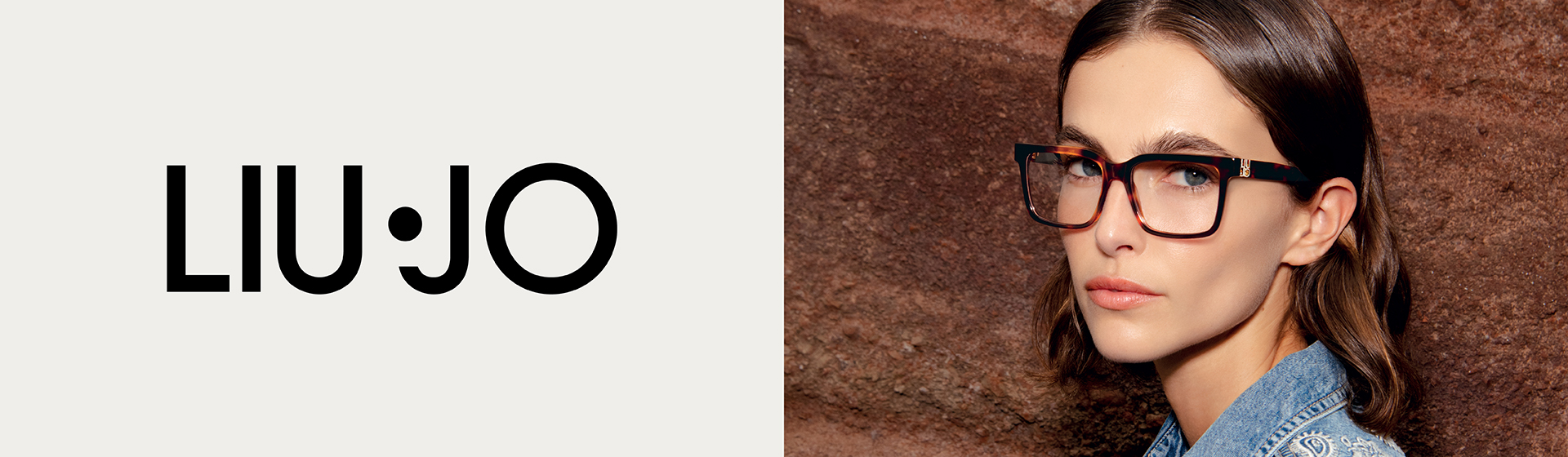 Una donna con gli occhiali, che indossa una giacca di jeans con motivi elaborati, è in piedi davanti a una parete marrone a texture. Si vede il logo Liu Jo.