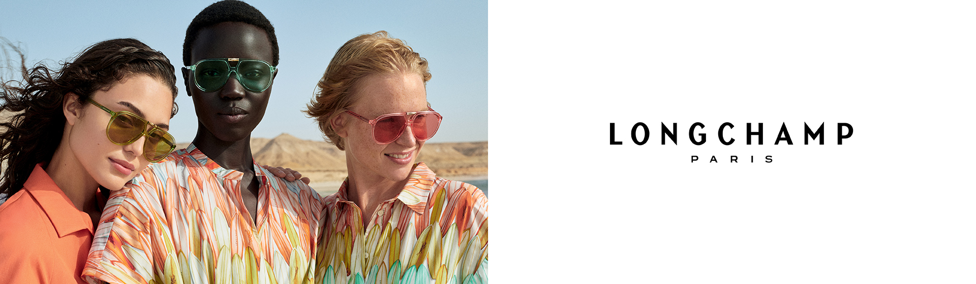Drei Models in bunten Kleidern und mit Sonnenbrillen stehen dicht beieinander und lächeln vor einer Wüstenkulisse. Auf dem Bild ist das Longchamp-Logo zu sehen.