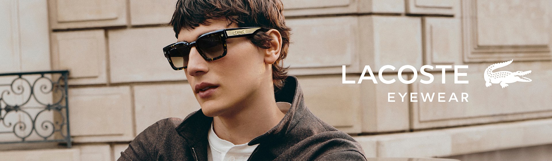 Osoba w ciemnych okularach przeciwsłonecznych i brązowej kurtce z logo Lacoste pozuje przed kamiennym murem na świeżym powietrzu. Widoczne jest logo Lacoste Eyewear.