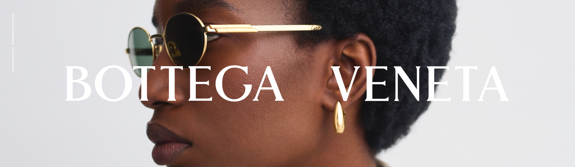 Person mit runder Sonnenbrille und goldenen Creolen, gekleidet in eine beige Jacke mit großem Kragen, mit dem Schriftzug „Bottega Veneta”.