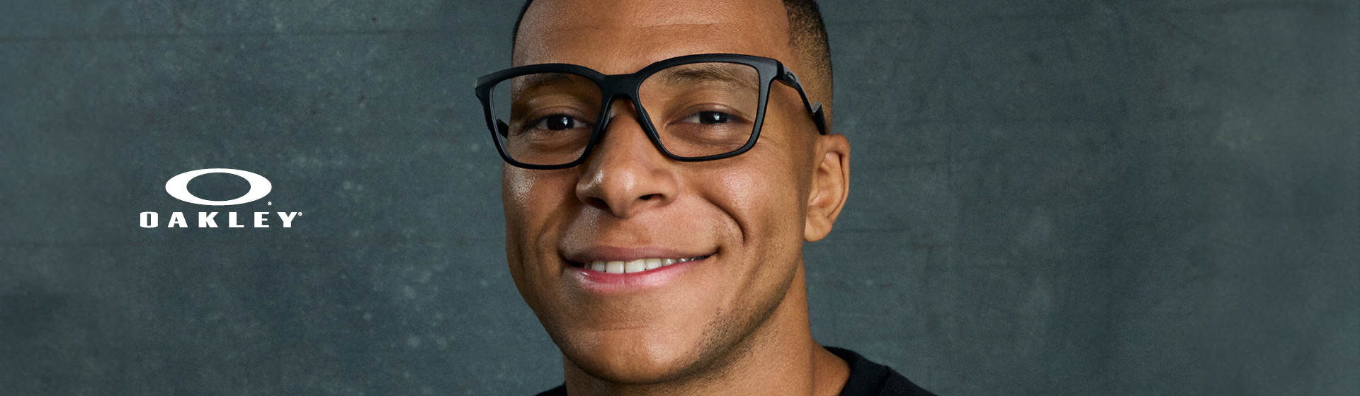 Osoba s černými brýlemi a černou košilí, usmívající se před strukturovaným šedým pozadím s logem Oakley.