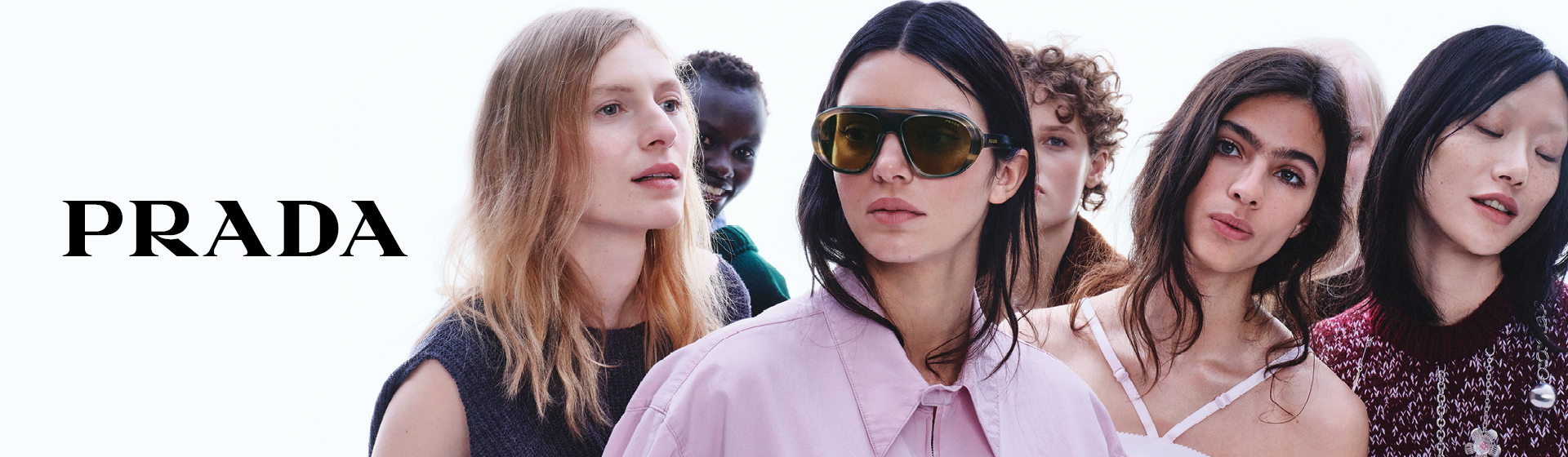 Eine Gruppe bunt und modisch gekleideter Personen posiert gemeinsam. Eine Person trägt eine große Sonnenbrille. In der unteren rechten Ecke ist „PRADA“ zu sehen.