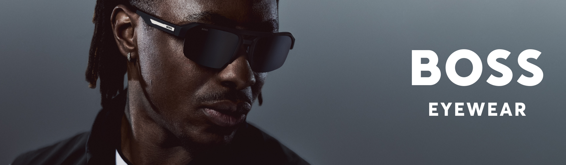 Una persona che indossa un paio di occhiali da sole alla moda con la scritta “BOSS EYEWEAR” sul lato destro, su uno sfondo scuro ed elegante.