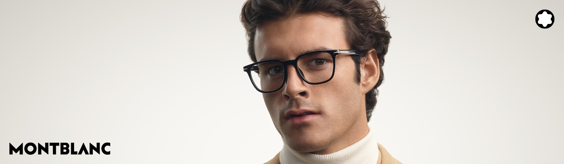 Un uomo con i capelli ricci e gli occhiali, che indossa un cappotto beige e un maglione a collo alto, davanti a uno sfondo neutro. È visibile il logo Montblanc.