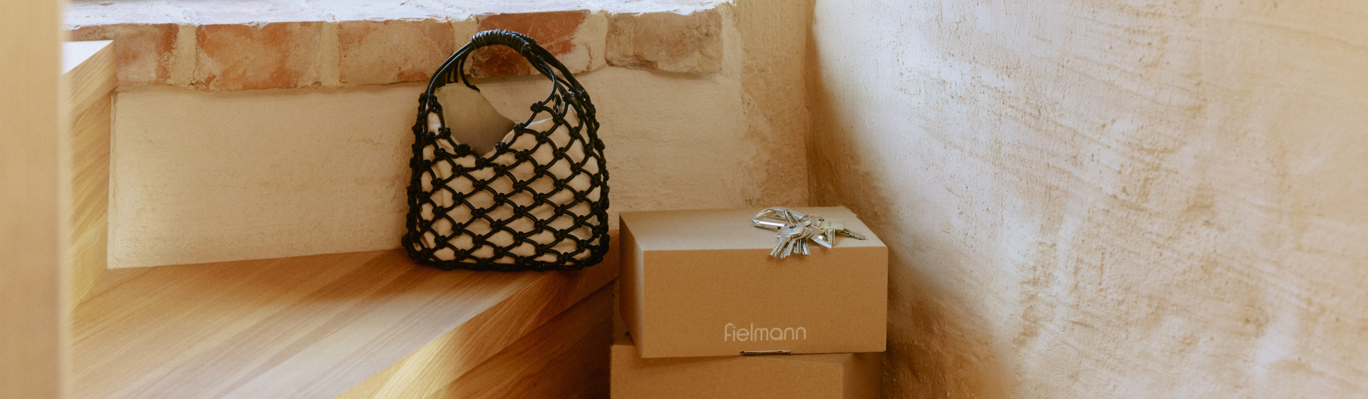 Gewebte schwarze Netztasche auf Holzstufen neben zwei gestapelten Paketen von Fielmann. Auf den Paketboxen liegt ein Schlüsselbund, dahinter eine strukturierte Gipswand.