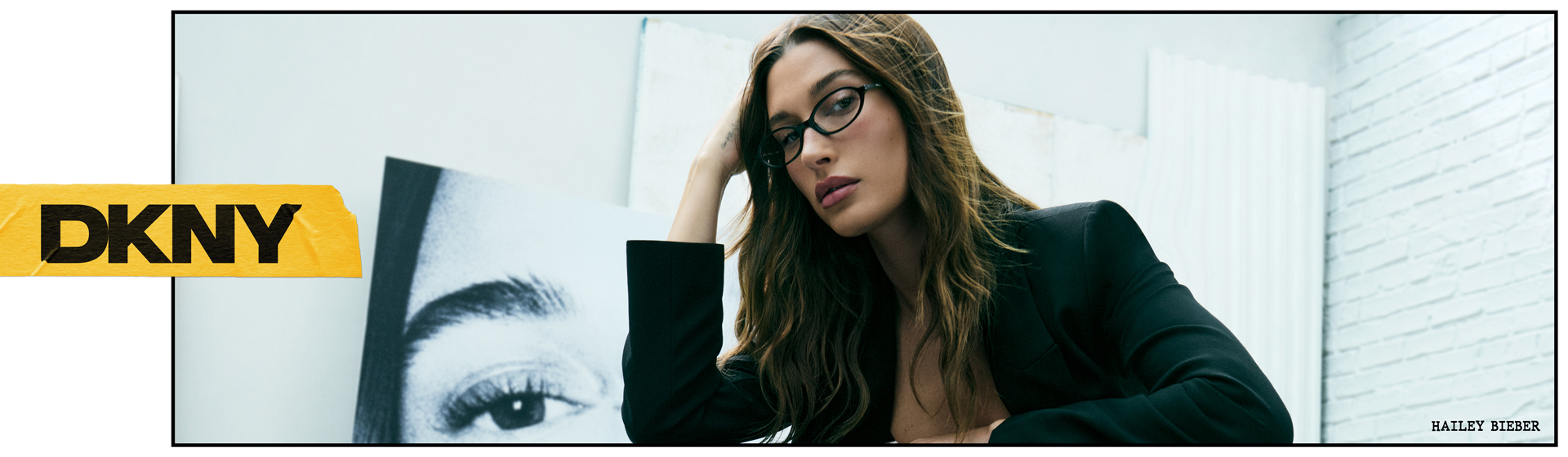 Une femme aux cheveux longs, portant des lunettes, pose vêtue d'un blazer noir, appuyée contre un bureau. On aperçoit le logo DKNY.
