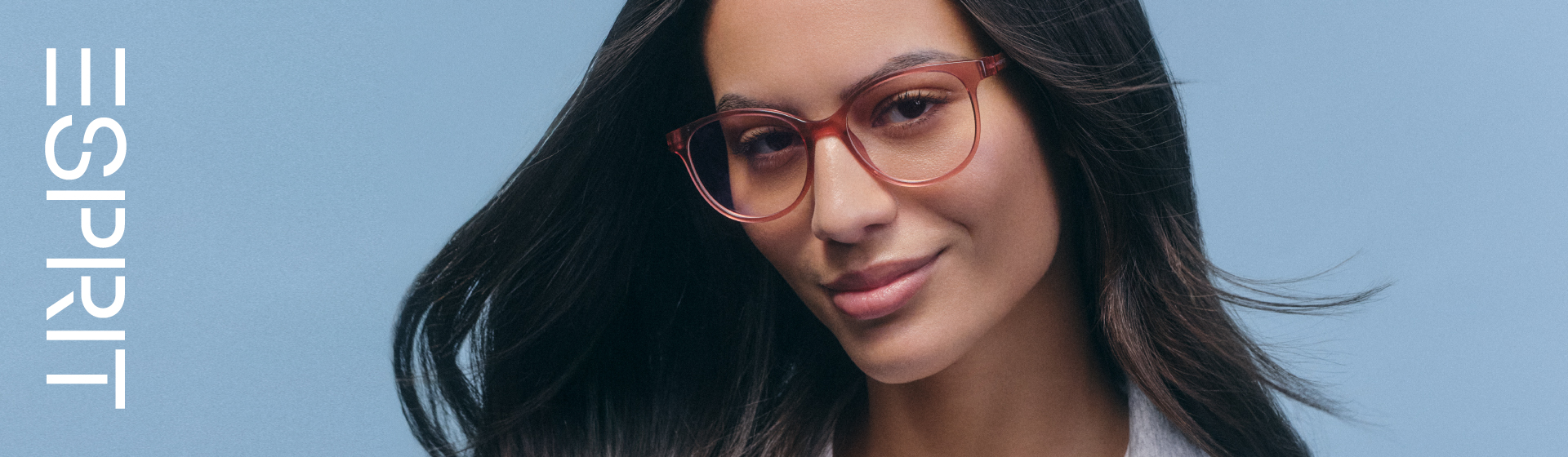 Žena s dlouhými vlasy a červenými brýlemi, která se lehce usmívá před modrým pozadím s logem „ESPRIT”.