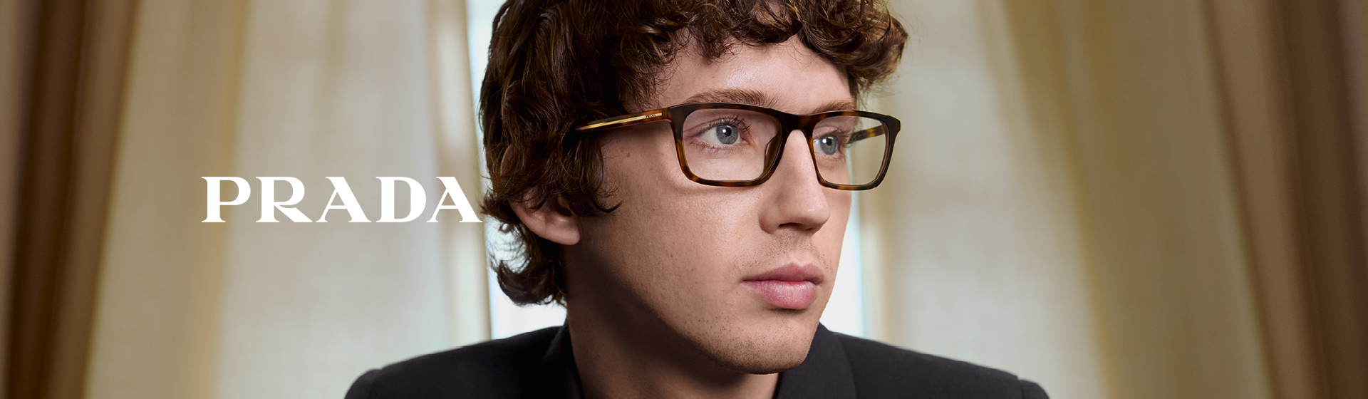 Eine Person mit Brille und Blazer, die zur Seite blickt; seitlich ist das Wort „PRADA“ zu sehen.