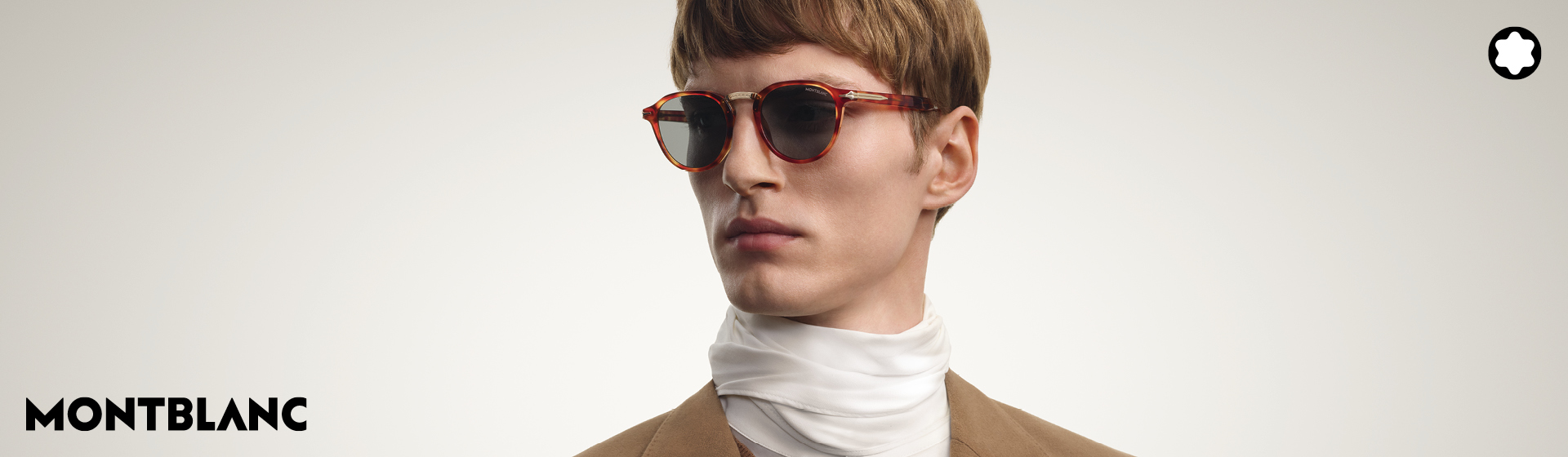 Eine Person mit Sonnenbrille und einer beigen Jacke sowie einem weißen Schal vor grauem Hintergrund. Montblanc-Logo in zu sehen.