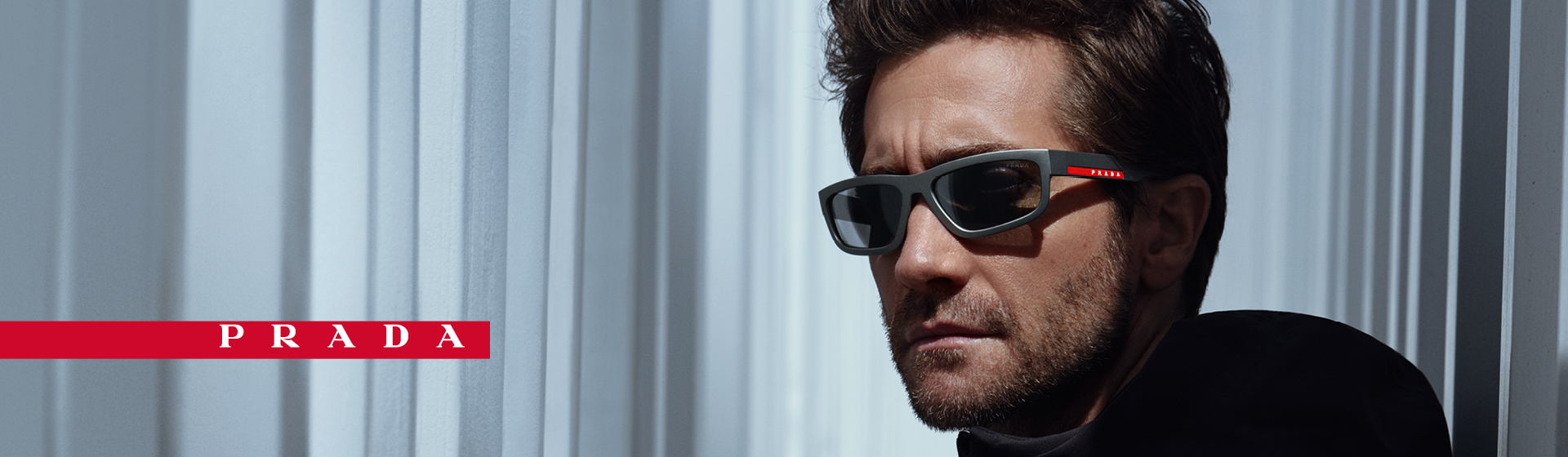 Un homme portant des lunettes de soleil et une veste noire pose avec un air sérieux devant un décor architectural moderne. On aperçoit un logo Prada.