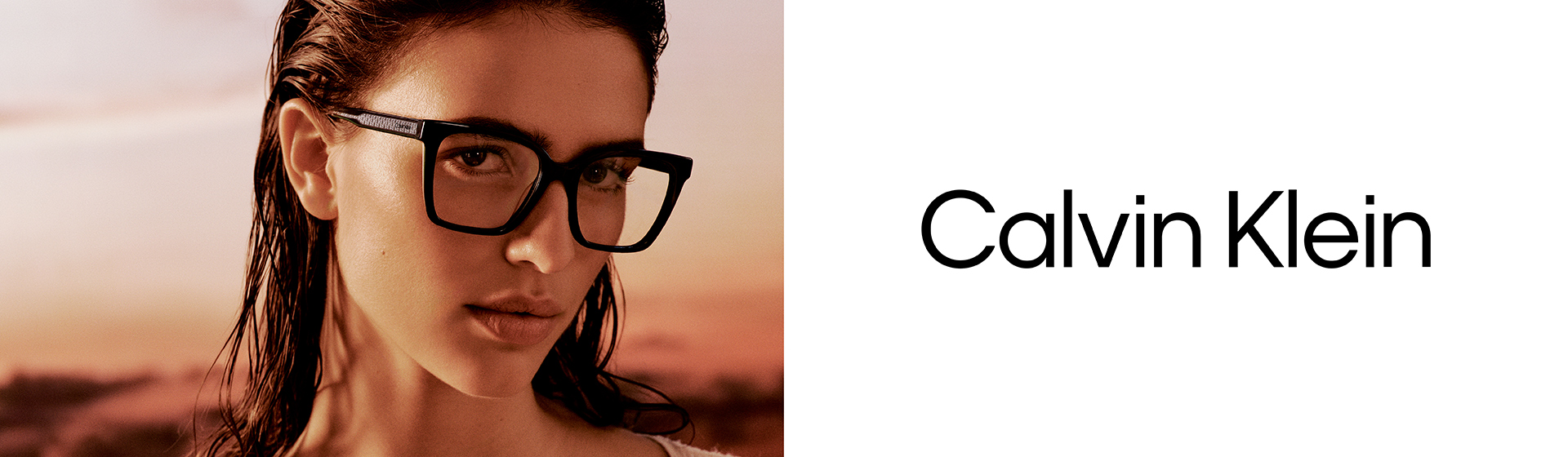 Osoba w ciemnych okularach i z mokrymi włosami, ubrana w białą bluzkę, pozuje na tle zachodu słońca. Widoczne logo Calvin Klein.