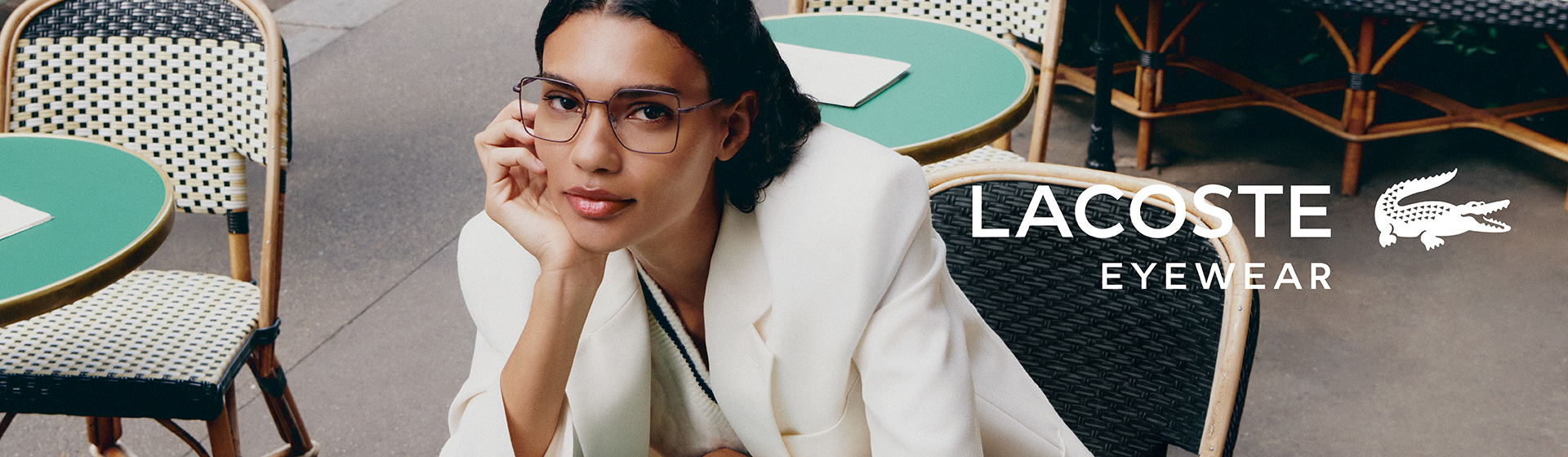 Osoba w białej marynarce i okularach, siedząca przy zielonym stole, z logo „Lacoste Eyewear”.