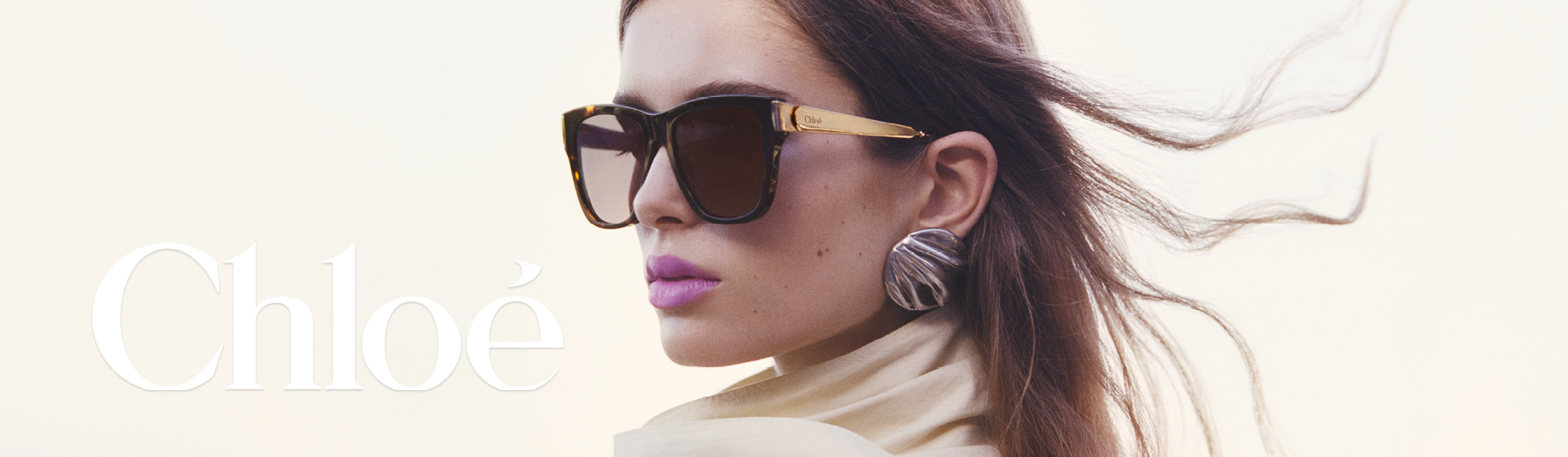 Žena s nadměrně velkými slunečními brýlemi a velkými strukturovanými náušnicemi, s vlasy rozcuchanými větrem, překrytá slovem „Chloé” v bílé barvě.