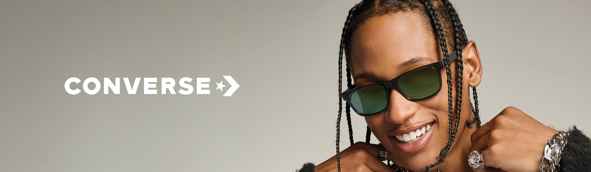 Personne aux cheveux tressés, portant des lunettes de soleil, une veste à motif léopard, un t-shirt imprimé et des bijoux, souriant devant un fond neutre. On aperçoit le logo Converse.