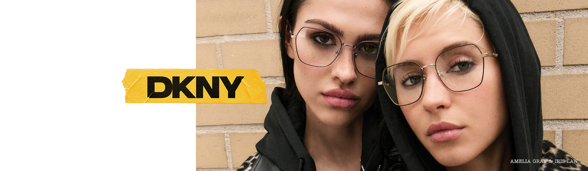 Zwei Personen in Kapuzenpullis und mit Brille posieren vor einer Backsteinmauer. Auf der linken Seite des Bildes ist das DKNY-Logo zu sehen.