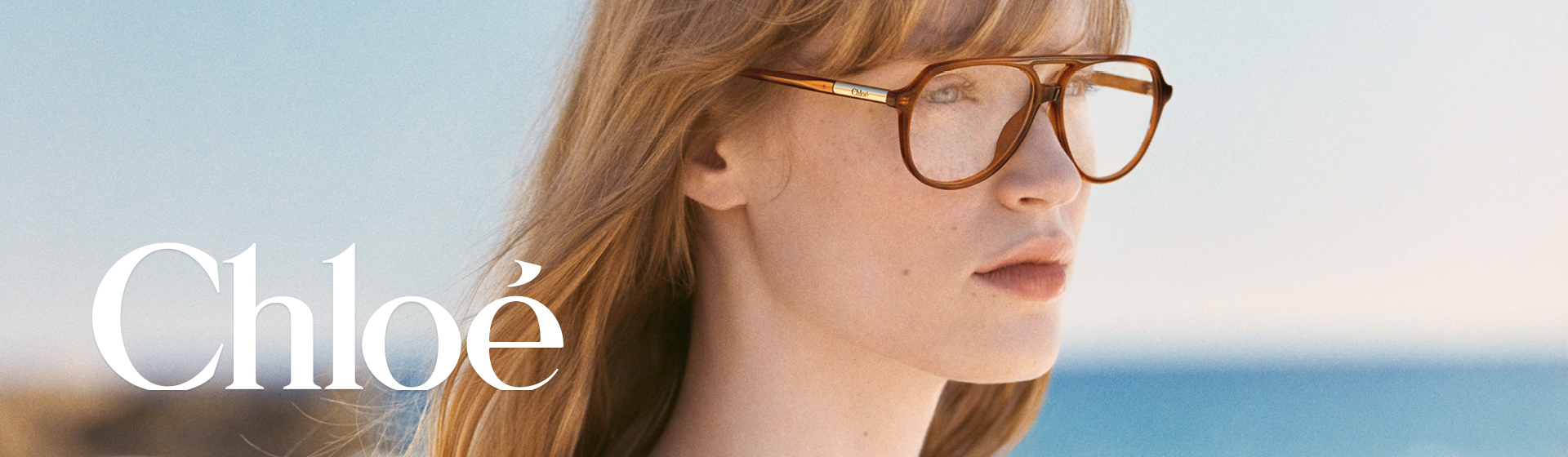 Žena s dlouhými vlasy a velkými brýlemi, má na sobě barevný vzorovaný top. Rozmazané pozadí pláže.