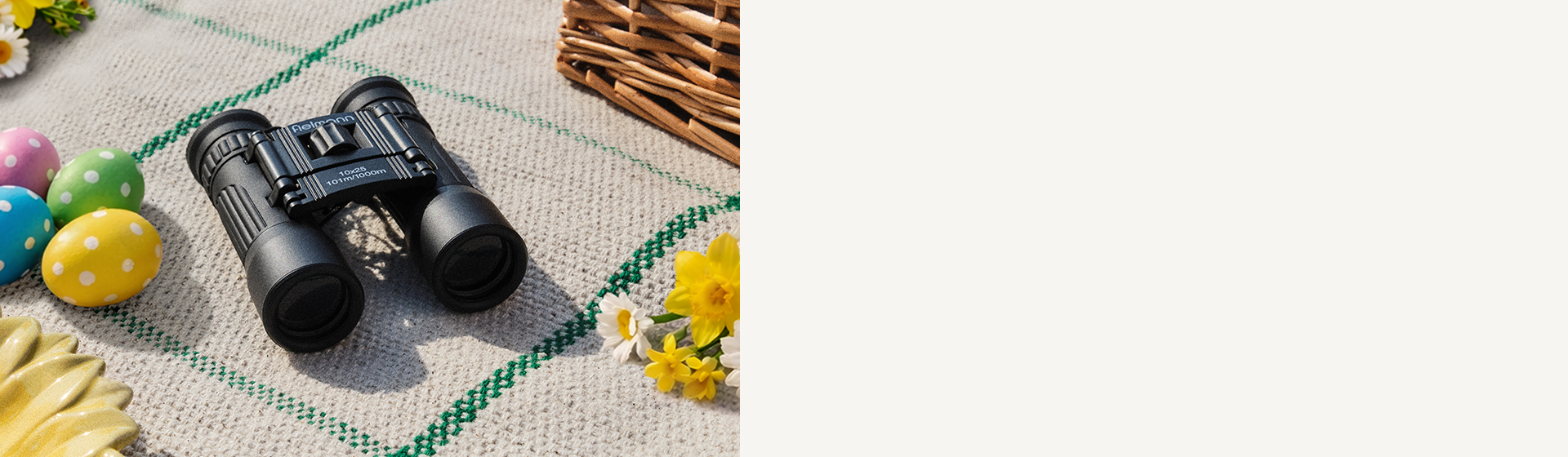 Ein Fernglas auf einer Picknickdecke, umgeben von bunten Ostereiern, gelben Blumen und einem Weidenkorb.