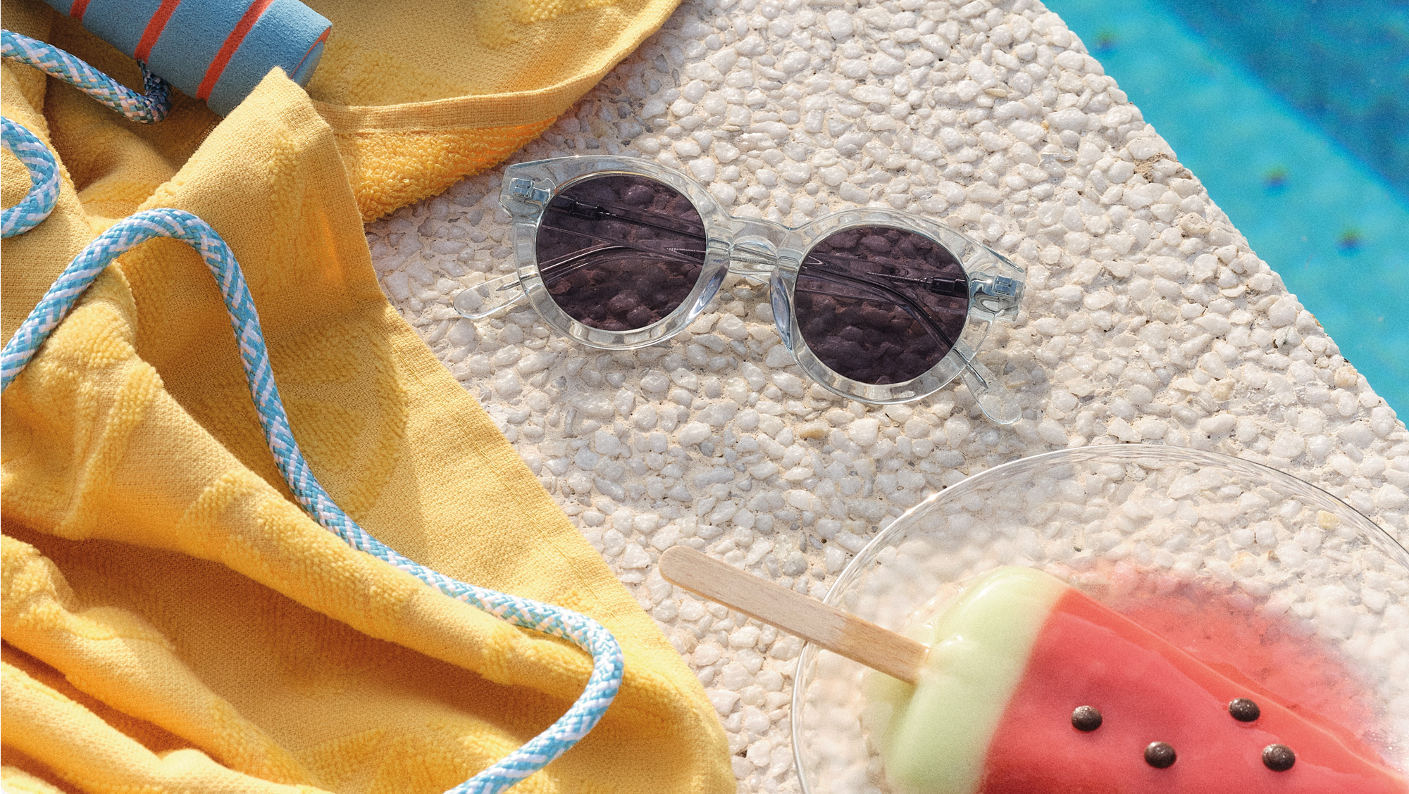 Sonnenbrille, ein gelbes Handtuch und ein Wassermelonen-Eis am Stiel liegen auf einer Steinoberfläche am Pool und vermitteln sommerliche Stimmung.