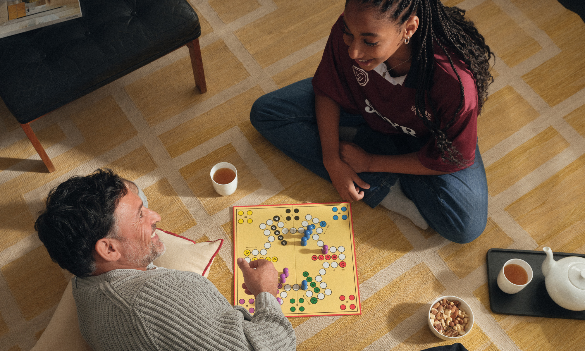 Mann mit Brille und Hörgerät spielt gemeinsam mit einer jungen Frau auf dem Boden ein Brettspiel. Auf dem Teppich stehen Tassen und eine Teekanne.