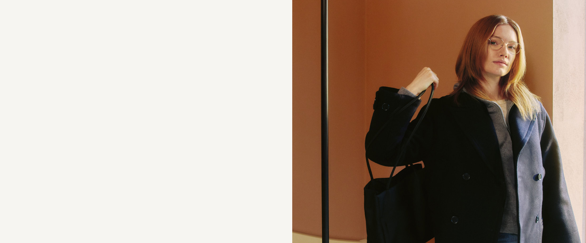 Eine Frau mit langen Haaren steht vor einem Spiegel, trägt einen dunklen Mantel und hält eine Tasche in der Hand, im Hintergrund ist eine Wand in warmen Farbtönen zu sehen.