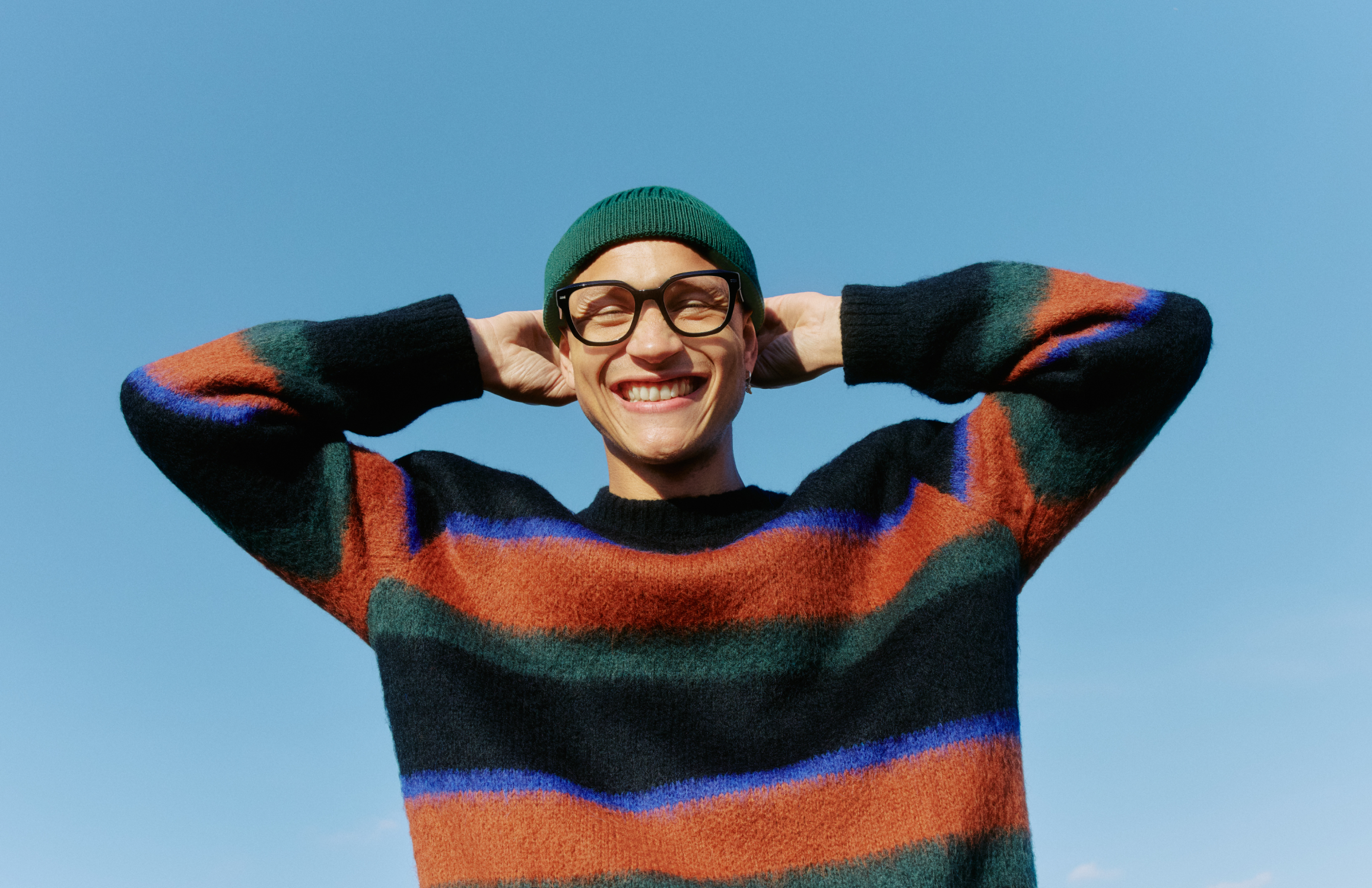Usmívající se muž s brýlemi a pruhovaným svetrem, se zeleným kloboukem, má ruce za hlavou na pozadí jasné modré oblohy.