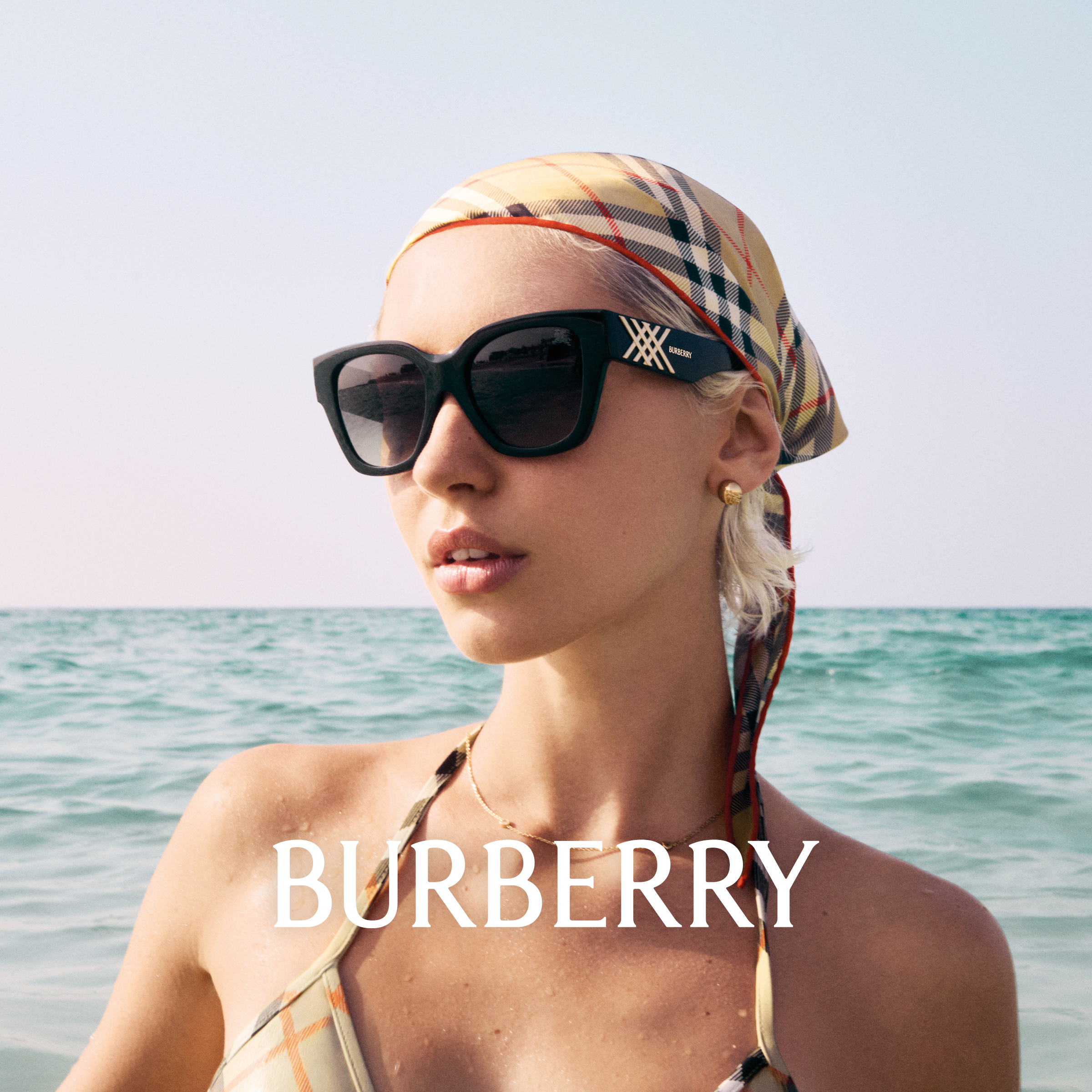 Eine Frau mit Sonnenbrille und Kopftuch mit Burberry-Muster steht am Strand, im Hintergrund das Meer.