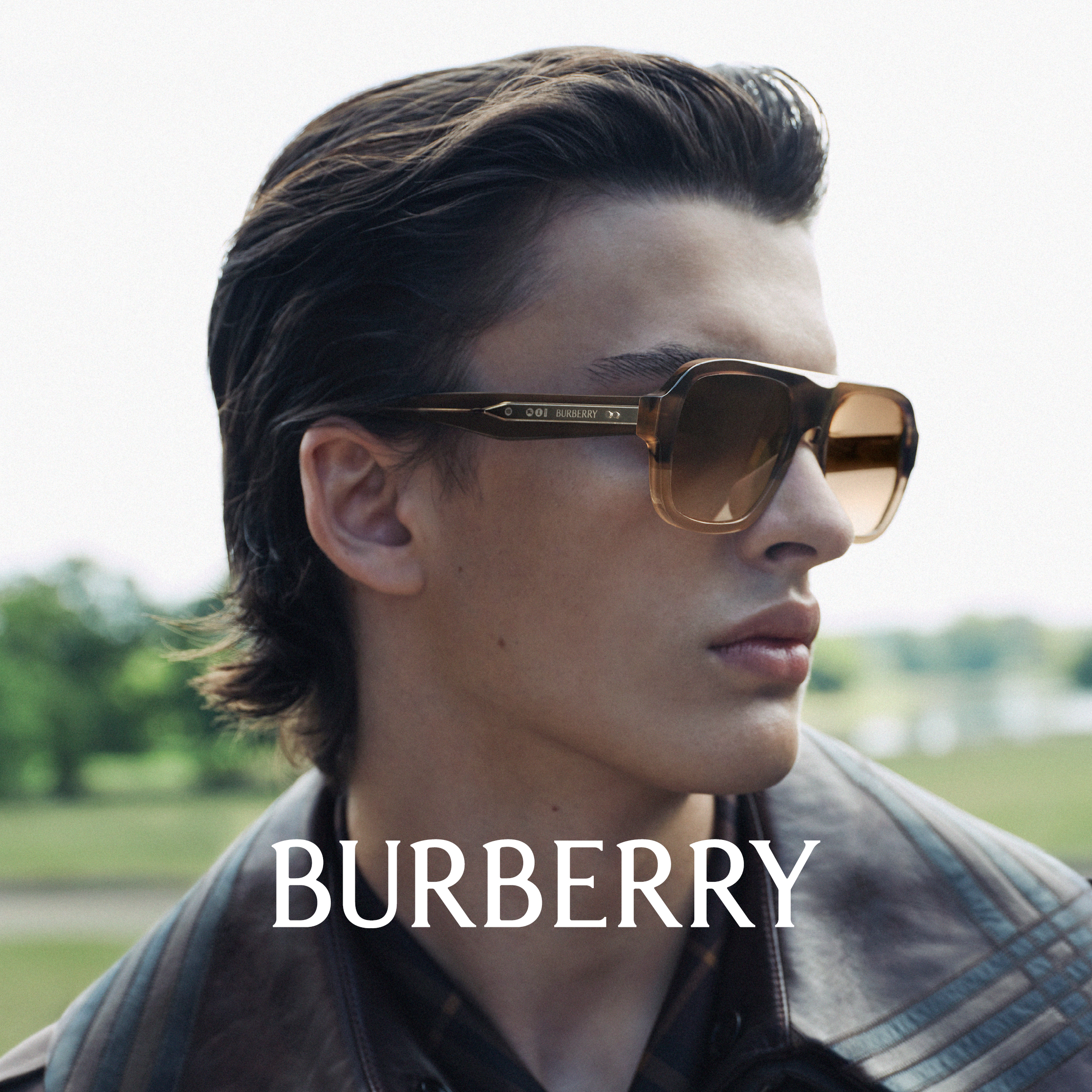 Uomo con grandi occhiali da sole e giacca di pelle, con la scritta “BURBERRY” sotto, all'aperto con uno sfondo verde sfocato.