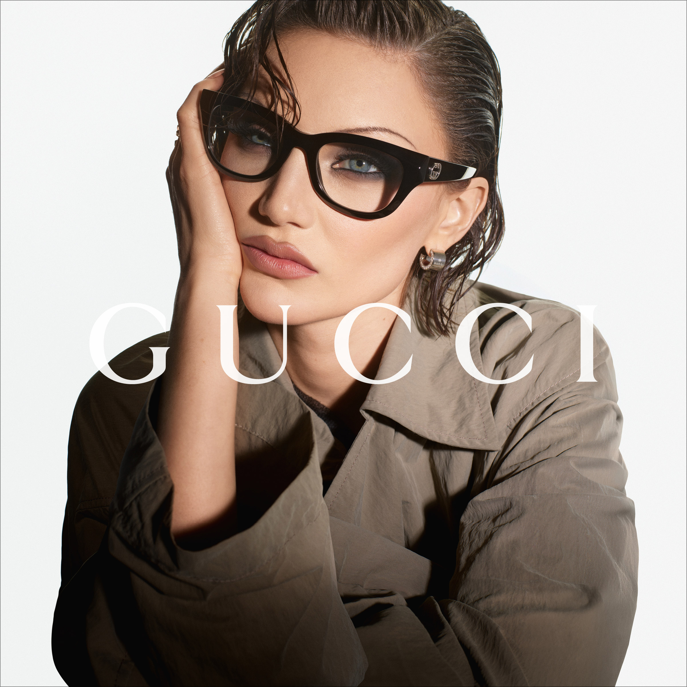 Una persona con i capelli lisciati all’indietro, che indossa occhiali con montatura scura e un trench beige e posa con una mano davanti al viso; sopra, la scritta “GUCCI”.