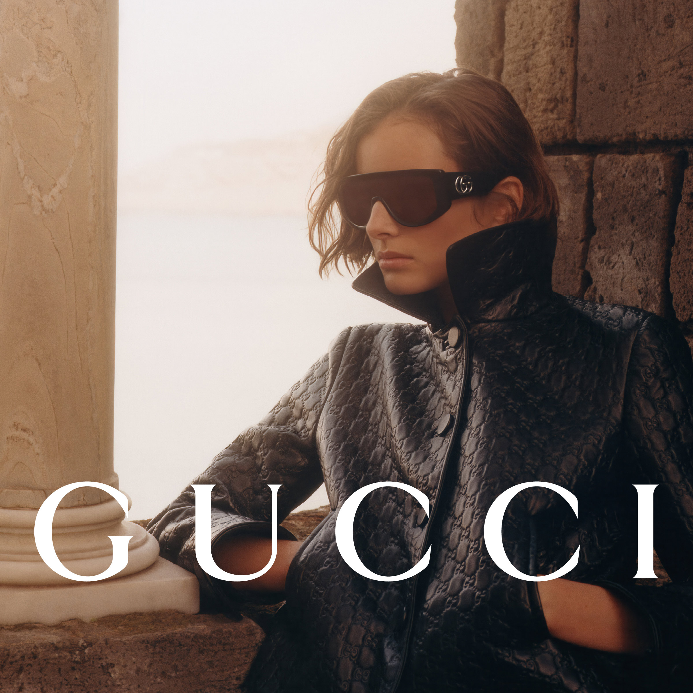 Donna con un cappotto nero strutturato e grandi occhiali da sole, appoggiata a una colonna di pietra con la scritta “GUCCI”.