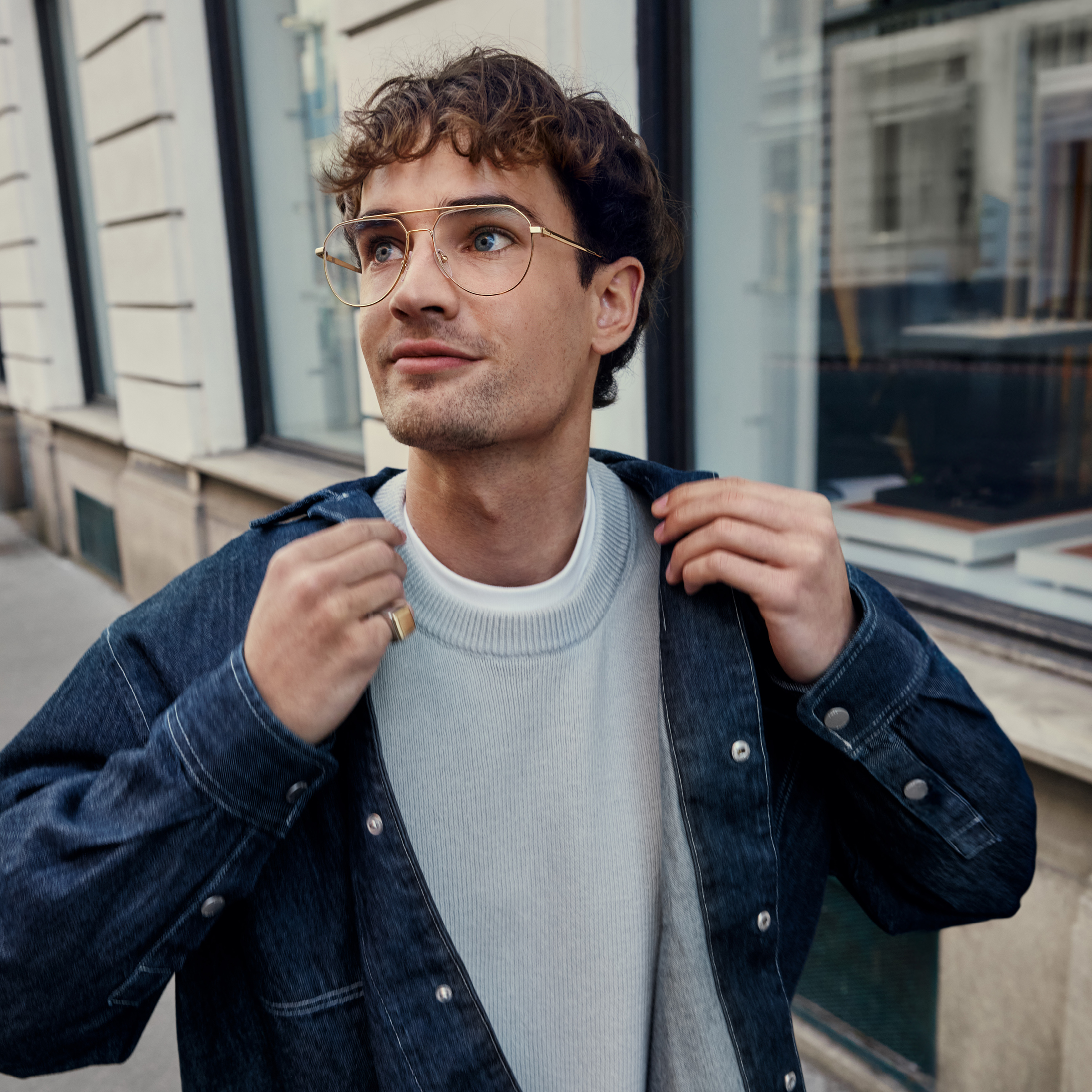 Ein Mann mit lockigem Haar und Brille steht auf einer Straße in der Stadt, zieht seine Jeansjacke über einem weißen Pullover zurecht und blickt nachdenklich nach oben.