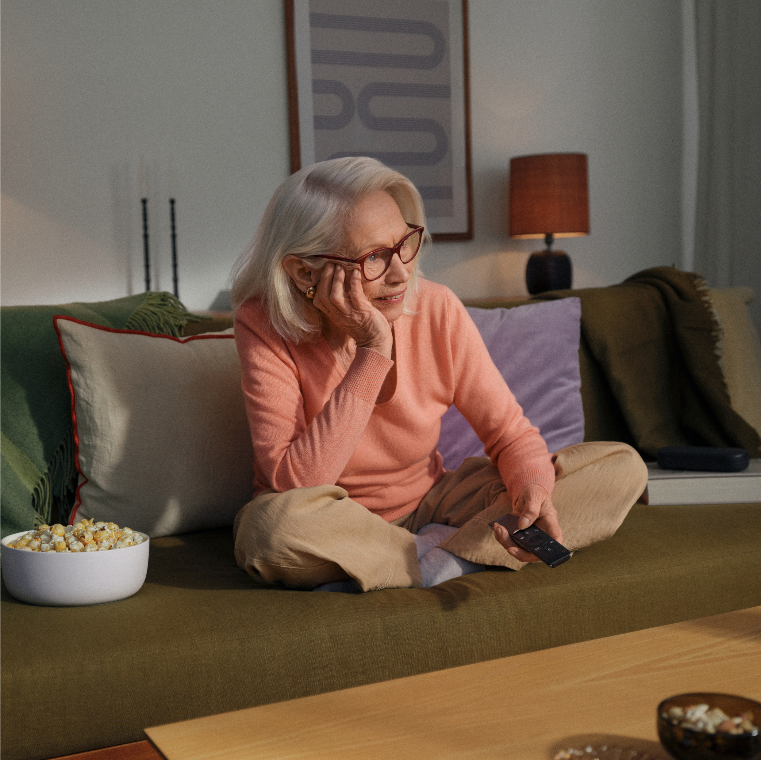Eine ältere Frau mit Brille, die einen pfirsichfarbenen Pullover trägt, sitzt mit gekreuzten Beinen auf einem Sofa, hält eine Fernbedienung in der Hand und hat eine Schüssel Popcorn neben sich stehen.