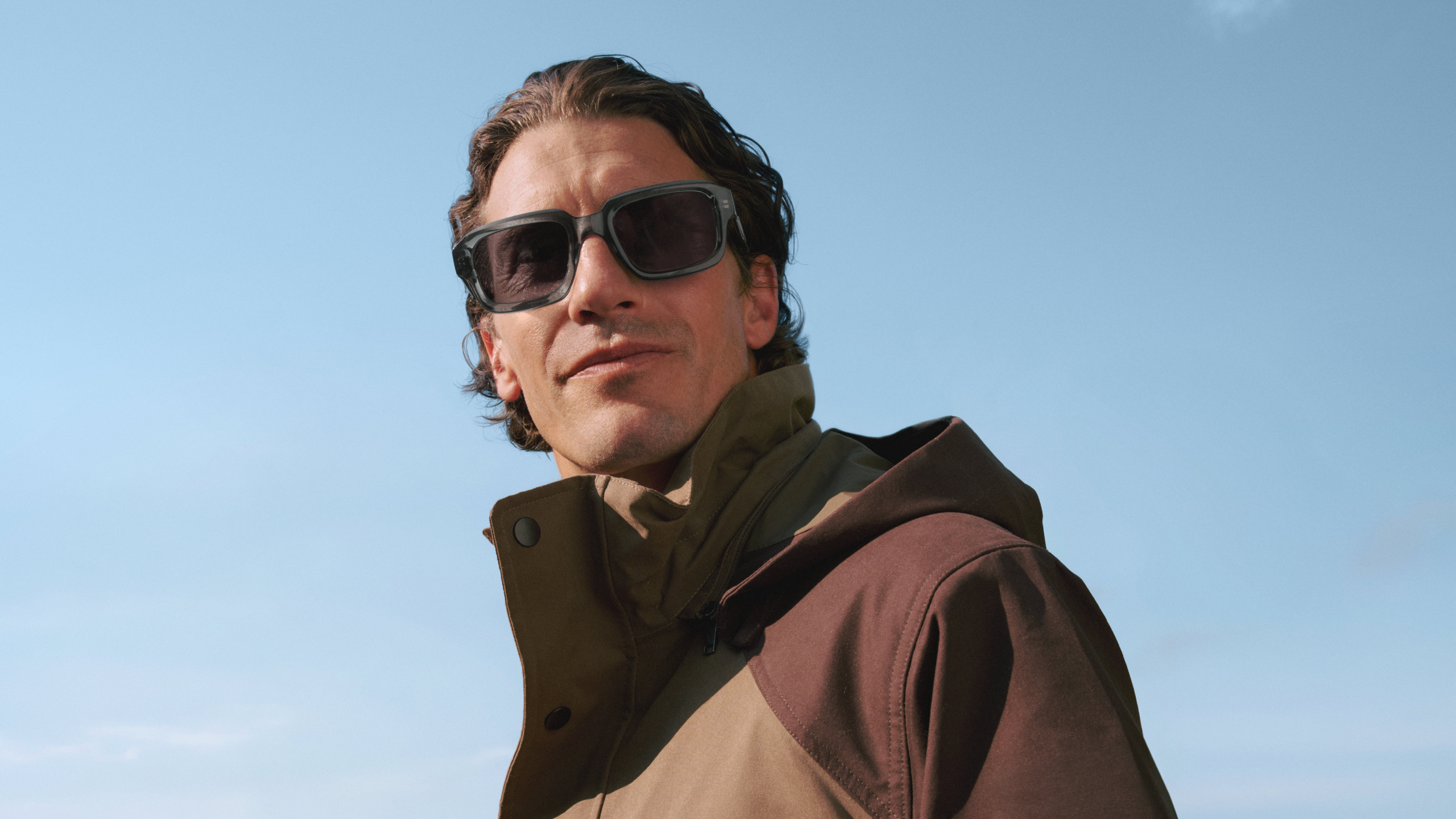 Ein Mann mit Sonnenbrille und brauner Jacke blickt vor einem strahlend blauen Himmel in die Ferne.
