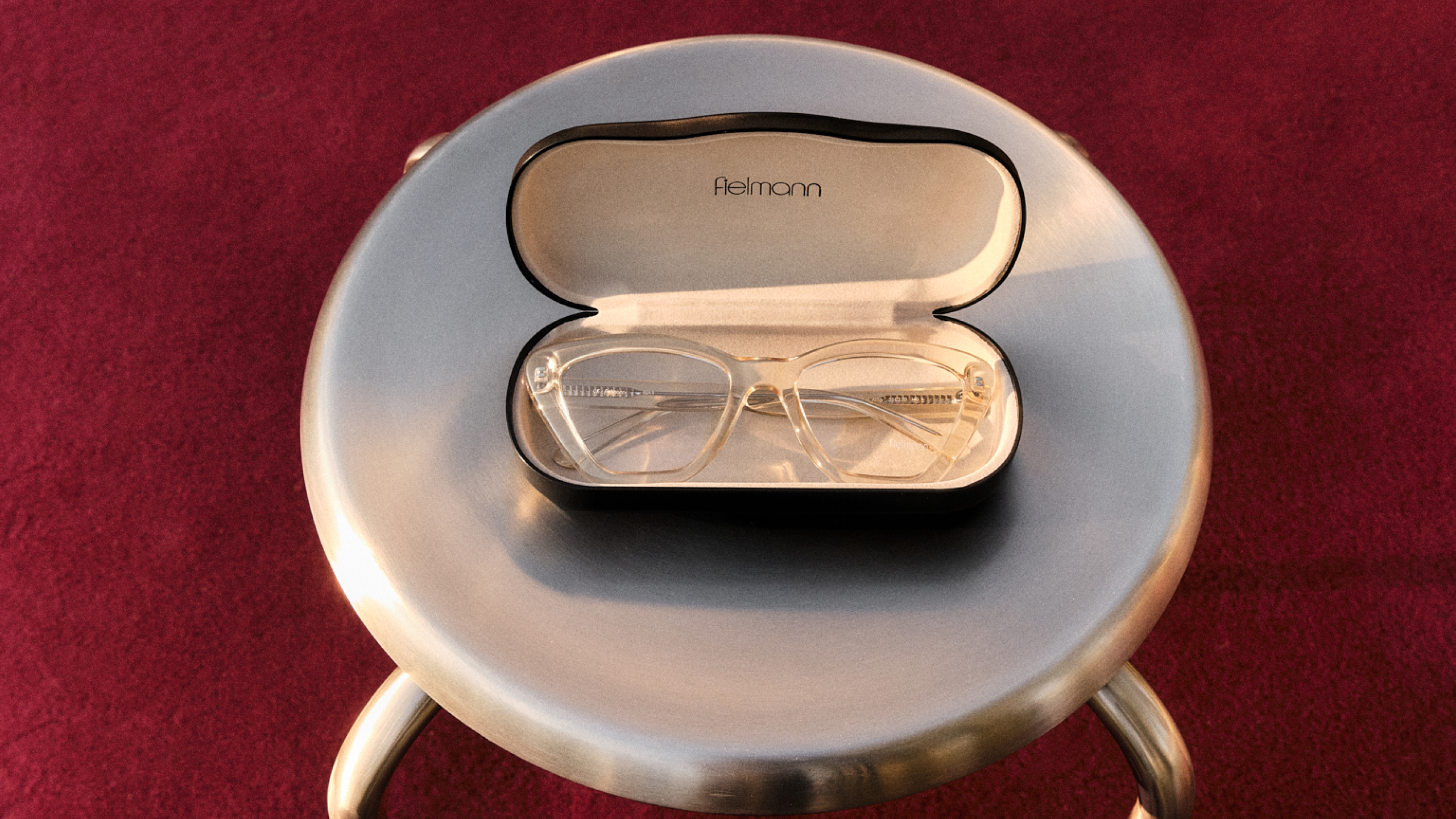 Durchsichtige Brille in einem geöffneten schwarzen Etui, platziert auf einem runden metallischen Hocker vor rotem Hintergrund.