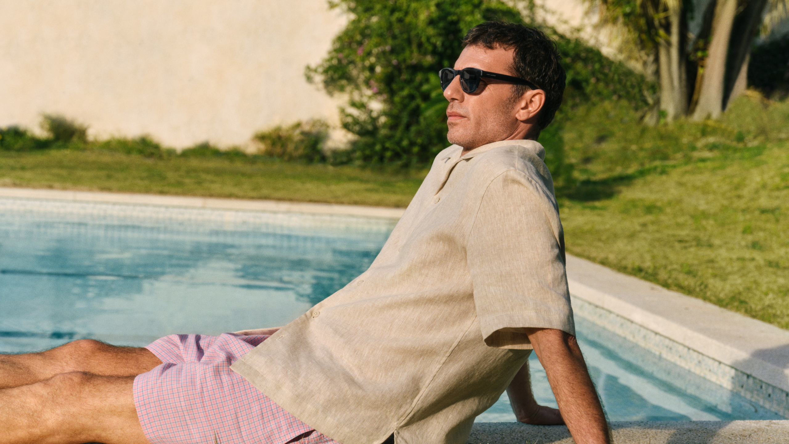 Un homme portant des lunettes de soleil, qui se détend au bord de la piscine, vêtu d'une chemise claire et d'un short rose, avec en arrière-plan un jardin ensoleillé.