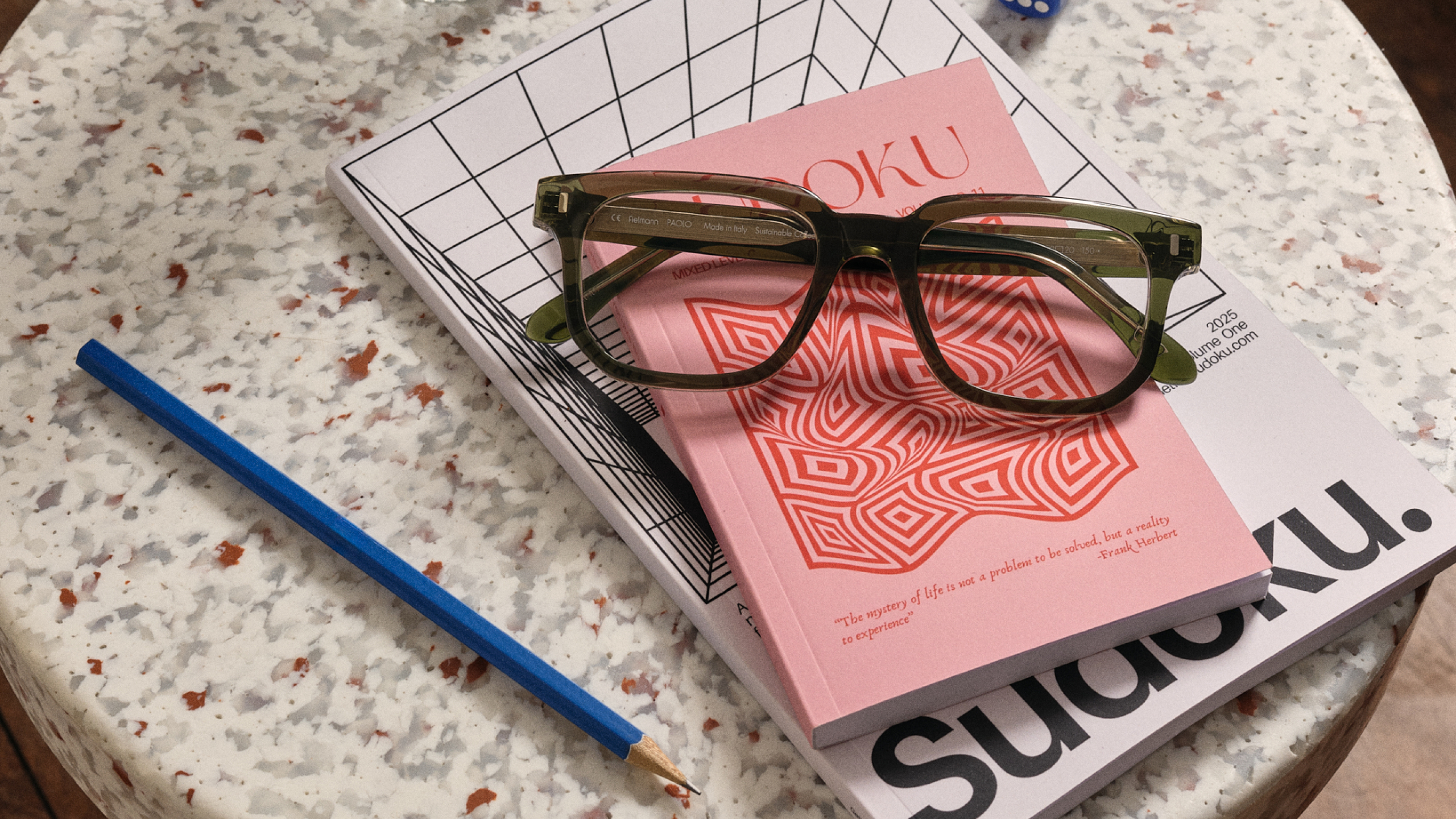 Eine Brille liegt auf rosa-weißen Sudoku-Rätselbüchern auf einem Tisch, daneben befindet sich ein blauer Bleistift.