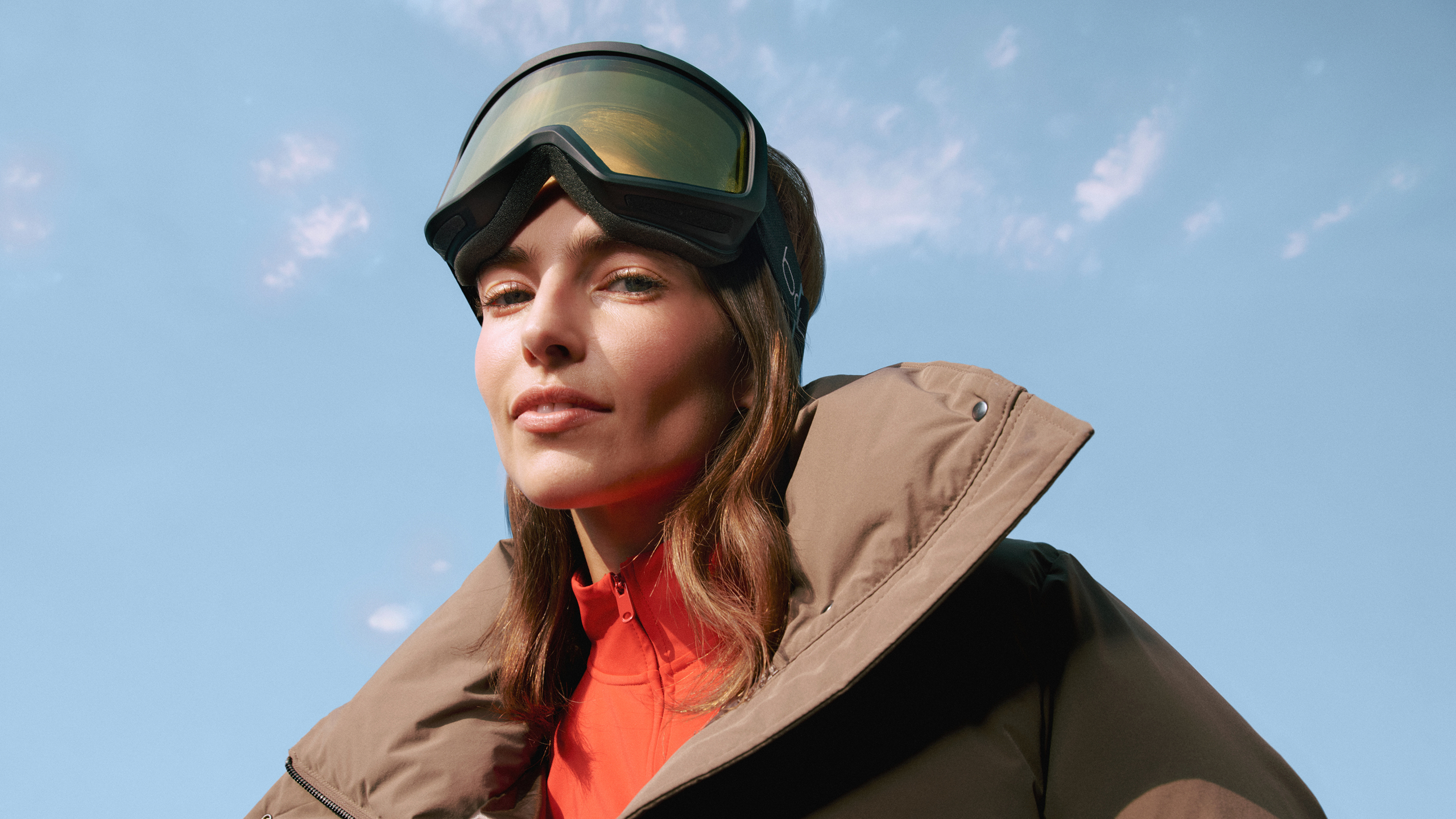 Eine Person in einer braunen Winterjacke und mit Skibrille blickt vor blauem Himmel selbstbewusst in die Kamera.